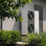 Eine moderne Wärmepumpe mit zwei großen runden Lüftungsöffnungen ist vor einem grauen Gebäude installiert, umgeben von grünen Büschen und Pflanzen.