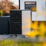Eine moderne Wärmepumpenanlage ist vor einem Haus auf einem Betonsockel in der Nähe einer Rasenfläche installiert. Im Hintergrund sind ein Fenster und ein Teil der Hauswand zu sehen, im Vordergrund ist etwas unscharfes gelbes Laub zu erkennen.
