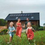 Eine lächelnde vierköpfige Familie, zwei Erwachsene und zwei Kinder, rennt und spielt auf einem Feld mit Gras und Wildblumen vor einem Haus mit Sonnenkollektoren auf dem Dach. Die Kinder halten Blumen und sehen glücklich aus.