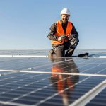 Ein Arbeiter mit weißem Schutzhelm und orangefarbener Sicherheitsweste kniet auf einem Dach neben Solarpaneelen, hält eine Bohrmaschine in der Hand und lächelt in den strahlend blauen Himmel.