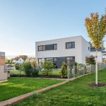 Ein modernes, zweistöckiges weißes Haus mit großen Fenstern steht in einem ordentlich angelegten Garten mit grünem Gras, Sträuchern und einem Baum. Ein Maschendrahtzaun trennt das Grundstück von den Nachbarhäusern im Hintergrund unter einem klaren blauen Himmel.