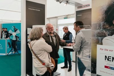 Ein Mann und eine Frau sprechen mit einem Aussteller an einem Messestand. Ein weiterer Mann steht in der Nähe, während im Hintergrund Messestände und Displays mit Branding zu sehen sind.