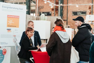 Eine Frau spricht mit drei Personen an einem Informationsstand mit der Aufschrift Energieberatung Saar in einer Veranstaltungshalle. Ein Mann steht in der Nähe und im Hintergrund ist ein Plakat mit Informationen zur Energieberatung zu sehen.