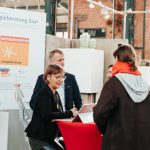 Eine Frau mit Brille sitzt an einem Tisch, benutzt einen Laptop und spricht mit einer Besucherin mit rotem Halstuch. Zwei Personen stehen hinter ihr in der Nähe einer Informationstafel der "Energieberatung Saar" bei einer Indoor-Veranstaltung.
