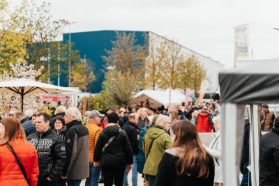 Eine große Menschenmenge in Jacken und Mänteln geht über einen Markt mit Zelten und Ständen im Freien. Im Hintergrund sind Bäume und ein blaues Gebäude bei bedecktem Himmel zu sehen.