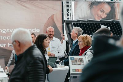 Eine Gruppe von Menschen tauscht sich aus und knüpft Kontakte auf einer belebten Indoor-Veranstaltung oder Messe, wobei im Hintergrund Auslagen, Informationsstände und Sonnenkollektoren zu sehen sind. Einige Teilnehmer lächeln und unterhalten sich.