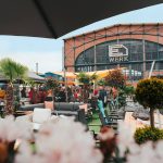 Vor einem großen Backsteingebäude mit der Aufschrift WERK versammeln sich Menschen auf einem Sitzplatz im Freien mit Palmen und bunten Sonnenschirmen. Die Szene ist lebendig und festlich, mit verschiedenen Möbeln für den Außenbereich.