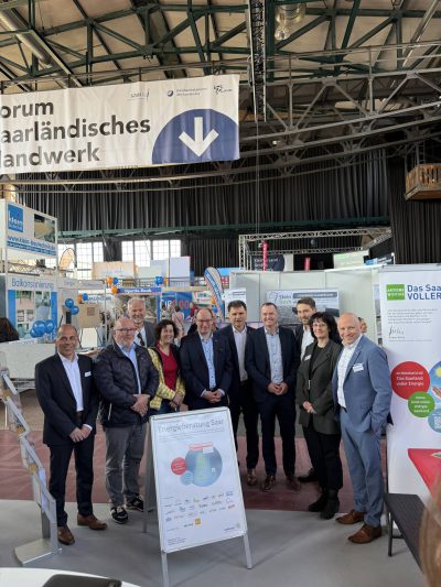 Eine Gruppe von Personen in Geschäftskleidung posiert für ein Foto auf einer Hallenausstellung. Hinter ihnen sind Informationsstände, Banner und ein großes Schild mit der Aufschrift "Forum saarländisches Handwerk" zu sehen.