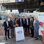 Eine Gruppe von zwölf Personen, gekleidet in Geschäftskleidung, steht in einem Innenraum auf einer Ausstellung oder Messe. Sie posieren gemeinsam hinter einer Schautafel mit Informationsgrafiken, wobei im Hintergrund Stände und Banner zu sehen sind.