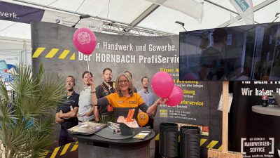 Eine Frau in einer orange-grauen Uniform lächelt, während sie zwei rosa Luftballons an einem Hornbach-Stand hält. Hinter ihr befindet sich eine Auslage mit Werkzeugen, Flyern, schwarzen Eimern und einem Poster mit Arbeitern. Auch ein Fernseher und eine Anlage sind zu sehen.