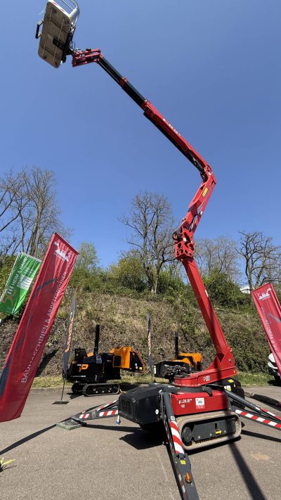 Ein roter Spinnenaufzug mit ausgefahrenem Auslegerarm und Korb ragt hoch über den Boden; in der Nähe stehen Baumaschinen und Banner, im Hintergrund Bäume und ein kleiner Hügel.