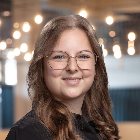 Eine junge Frau mit langen, gewellten braunen Haaren und einer Brille lächelt in die Kamera. Sie trägt ein schwarzes Oberteil, und im Hintergrund sind Hängelampen und eine sanft verschwommene Innenraumkulisse zu sehen.