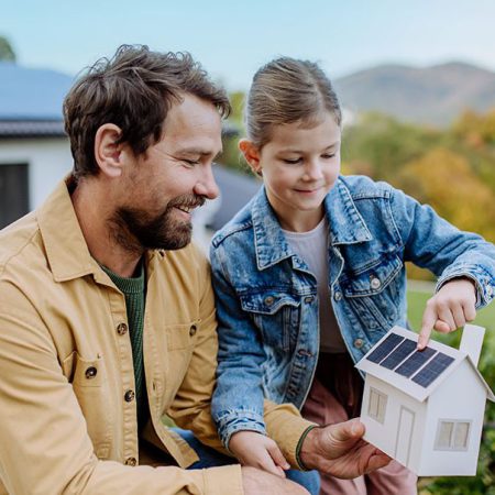 Ein Mann und ein junges Mädchen lächeln im Freien, während das Mädchen auf die Solarzellen an einem kleinen Modellhaus zeigt, das sie zusammenhalten.
