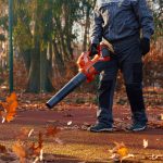 Eine Person in Arbeitskleidung verwendet einen orangefarbenen Laubbläser, um herabgefallene Herbstblätter von einem von Bäumen umgebenen Sportplatz im Freien zu entfernen. Die Szene spielt bei Tageslicht, die Blätter sind verstreut und wirbeln durch die Luft.