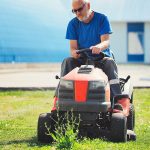 Ein älterer Mann mit Sonnenbrille und blauem Hemd fährt auf einem roten Rasenmäher und mäht an einem sonnigen Tag Gras. Im Hintergrund ist ein verschwommenes Gebäude zu sehen.