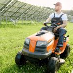 Ein Mann in einem Arbeitsoverall und mit einer Mütze bedient einen orangefarbenen Aufsitzrasenmäher auf einer Wiese in der Nähe großer Solarzellenfelder unter einem sonnigen Himmel.
