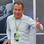 Ein Mann mit einem hellgrauen Hemd und einem grünen Schlüsselband lächelt und zeigt den Daumen nach oben. Er steht in einem Innenraum vor einer Kulisse mit Autositzen und einem Sternenhintergrund.