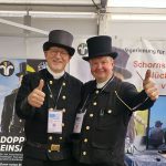Zwei lächelnde Männer in traditionellen schwarzen Schornsteinfegeruniformen und Zylinderhüten stehen nebeneinander und zeigen die Daumen nach oben. Sie tragen Veranstaltungsplaketten und stehen vor einem Stand bei einer Indoor-Veranstaltung.