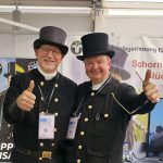 Zwei lächelnde Männer in traditionellen Schornsteinfegeruniformen und Zylinderhüten stehen nebeneinander und zeigen die Daumen nach oben. Sie befinden sich in einem Innenraum auf einer Veranstaltung, mit Namensschildern und Informationsbannern hinter ihnen.
