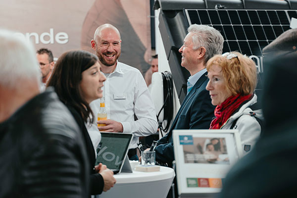 Eine Gruppe von Personen unterhält sich bei einer Veranstaltung in einem Gebäude. Drei Männer und zwei Frauen unterhalten sich in der Nähe von Ausstellungstischen mit Broschüren, Laptops und Solarzellen im Hintergrund.