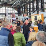 Eine große Gruppe von Menschen geht umher und versammelt sich auf einer überdachten Ausstellung oder Messe mit verschiedenen Ständen und Displays. Die Menge scheint engagiert zu sein, und der Veranstaltungsort verfügt über hohe Decken und große Fenster.