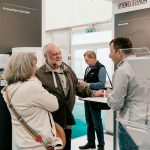 Drei Personen unterhalten sich auf einer Messe zum Thema erneuerbare Energien mit einem Standvertreter, während im Hintergrund Informationsdisplays und Produktbroschüren zu sehen sind.