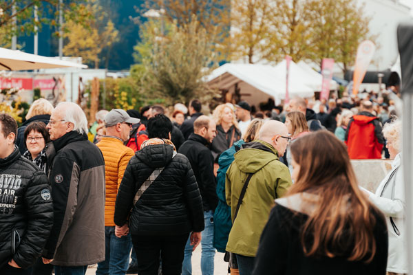 Eine große, in Jacken gekleidete Menschenmenge geht und steht an einem bewölkten Tag im Freien bei einer geschäftigen Veranstaltung mit Zelten und Bäumen im Hintergrund.