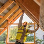 Ein Bauarbeiter, der eine gelbe Sicherheitsweste und einen Helm trägt, prüft mit einer Wasserwaage die Ausrichtung eines hölzernen Dachstuhls in einem halbfertigen Haus.