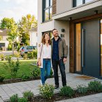 Ein lächelndes Paar steht vor einem modernen Haus und sieht glücklich aus. Die Frau hält den Arm des Mannes, und er hält die Schlüssel. Das Haus hat große Fenster, Pflanzen und einen gepflasterten Gehweg. Im Hintergrund sind andere Häuser und Bäume zu sehen.
