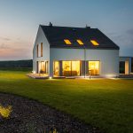 Ein modernes zweistöckiges Haus mit großen Fenstern, die in der Abenddämmerung warm leuchten, umgeben von einem gepflegten Rasen und einem kleinen Landschaftsgarten, vor einem ruhigen Abendhimmel.