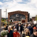 Vor einem großen Industriegebäude aus Backstein mit Glasfenstern, die mit WERK beschriftet sind, herrscht an einem sonnigen Tag reges Treiben. Die Menschen unterhalten sich, tragen Luftballons und kaufen an verschiedenen Ständen im Freien ein.