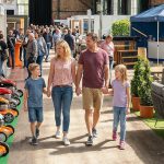 Eine vierköpfige Familie, zwei Erwachsene und zwei Kinder, geht Hand in Hand durch eine Halle mit Rasenmähern und anderen Ausstellungsstücken, während sich andere Personen im Hintergrund an den Ständen umsehen.