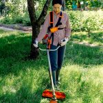 Eine Person mit Schutzhandschuhen und Stiefeln schneidet mit einem orange-weißen Fadentrimmer hohes Gras in einem Garten, während sie in der Nähe eines Baumes steht und von üppigem Grün umgeben ist.
