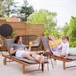 Ein Mann, eine Frau und ein junges Mädchen entspannen sich auf Liegestühlen an einem Pool, lächeln und halten Getränke in der Hand. Neben ihnen steht ein kleiner Tisch mit Obst, und im Hintergrund sind Bäume und ein Holzzaun zu sehen.