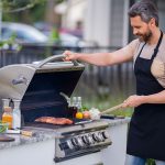 Ein Mann mit einer schwarzen Schürze grillt Fleisch auf einem Gasgrill im Freien. Mit der einen Hand hebt er den Deckel an, in der anderen hält er einen Spachtel. Auf der Theke neben ihm liegen verschiedene Gewürze und Gemüse.