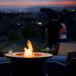 Ein Paar steht Arm in Arm und betrachtet einen malerischen Sonnenuntergang über einer Stadt. Im Vordergrund sorgen zwei Stühle und ein Tisch mit Feuerstelle und zwei Gläsern Wein für ein gemütliches Ambiente im Freien.