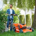 Ein Mann in Freizeitkleidung schiebt einen orangefarbenen Rasenmäher und mäht Gras in einem sonnigen, grünen Garten mit Bäumen und Sträuchern um ihn herum.