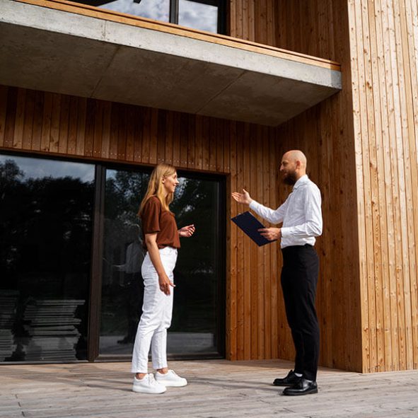 Ein Mann mit einem blauen Ordner unterhält sich mit einer Frau vor einem modernen Holzgebäude, beide stehen bei Tageslicht auf einem Holzdeck.