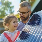 Ein Mann und ein junges Mädchen lächeln sich an, während sie an einem sonnigen Tag im Freien ein Solarmodul halten, mit Grünpflanzen und einem Gebäude im Hintergrund.