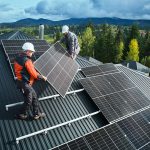 Zwei Arbeiter mit Schutzhelmen installieren bei teilweise bewölktem Himmel Solarmodule auf dem Dach eines Gebäudes, das von Bäumen und Bergen umgeben ist.