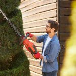 Ein Mann mit Schutzbrille und roten Handschuhen schneidet mit einer orangefarbenen elektrischen Heckenschere einen hohen Busch in einem Garten, der an einer Holzwand steht.
