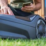 Eine Person benutzt ein Smartphone, während sie ihre Hand auf einen Roboter-Rasenmäher legt, der auf grünem Gras steht. Das große Rad und das schlanke Design des Mähers sind im Vordergrund zu sehen.