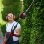 Eine Person, die einen Gehörschutz, Handschuhe und einen Arbeitsoverall trägt, schneidet mit einer elektrischen Heckenschere an einer langen Stange hohe grüne Sträucher, während sie im Freien in einem Garten oder Hof steht.