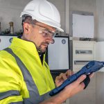 Ein männlicher Arbeiter in Warnweste, Schutzhelm und Schutzbrille verwendet ein robustes Tablet, während er in einem Gebäude in der Nähe von elektrischen Anlagen steht.
