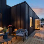 Moderne Dachterrasse in der Abenddämmerung mit gemütlichen Sitzgelegenheiten, Topfpflanzen und sanfter Beleuchtung. Schwarz getäfelte Außenwände und Holzterrassen schaffen einen stilvollen, einladenden Lebensraum im Freien.