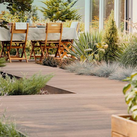 Ein hölzerner Gartenweg führt zu einem Esstisch mit Holzstühlen im Freien, umgeben von üppigen grünen Pflanzen und Sträuchern in der Nähe eines modernen Glasgebäudes.