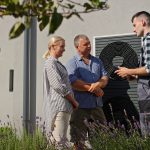 Ein Mann in Arbeitskleidung erklärt einem lächelnden Paar, das vor einem modernen Haus in der Nähe einer großen Wärmepumpenanlage steht, etwas, wobei Pflanzen und Lavendel im Vordergrund zu sehen sind.