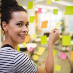 Eine Frau in einem gestreiften Hemd lächelt in die Kamera, während sie in einem hellen Büro bunte Haftnotizen an einer Glaswand anbringt.