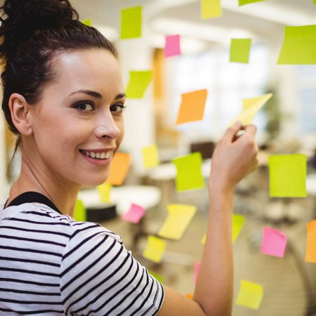 Eine Frau in einem gestreiften Hemd lächelt in die Kamera, während sie in einem hellen Büro bunte Haftnotizen an einer Glaswand anbringt.