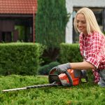 Eine Frau mit Brille, Handschuhen und einem rotkarierten Hemd schneidet in einem Garten eine grüne Hecke mit einer elektrischen Heckenschere. Im Hintergrund ist ein Haus mit einem roten Dach zu sehen.